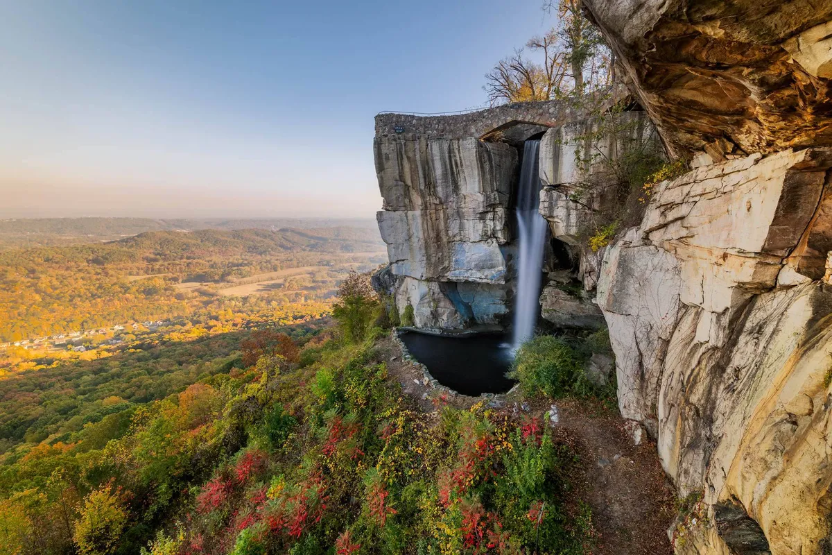 A breathtaking scene featuring a waterfall cascading over rock formations, surrounded by autumn foliage and a sweeping valley view. The atmosphere is peaceful and immersive, inviting exploration and relaxation in nature.