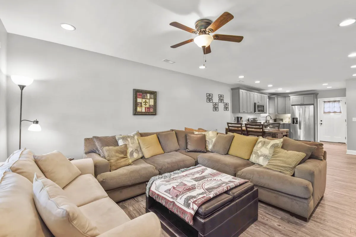 A spacious, open living room featuring a large sectional sofa and a coffee table with thoughtful decor. In the background, the adjoining kitchen area is visible, reinforcing the open-concept layout. The scene exudes a cozy, inviting atmosphere perfect for relaxing, hosting game nights, or gathering with friends and family.