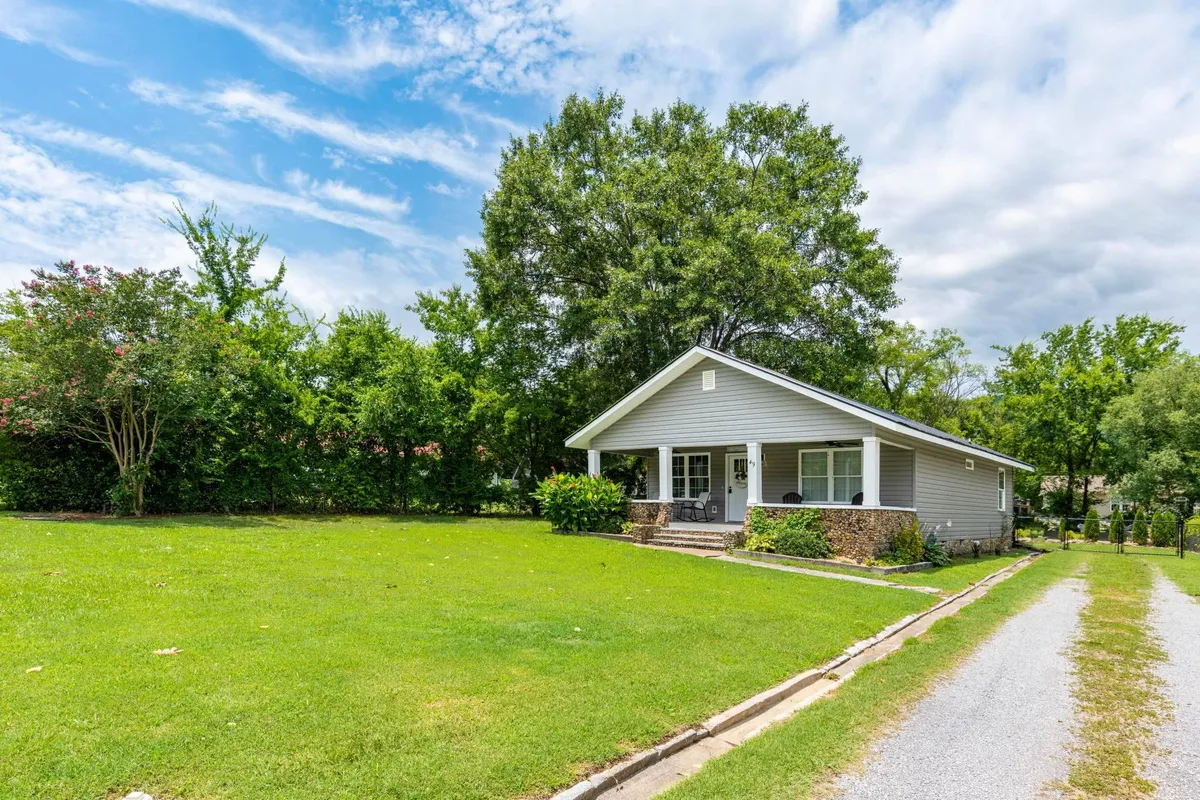 A welcoming view of the home’s front porch, the expansive lawn, and mature trees surrounding the property. The open green space is clearly visible, suggesting plenty of room for pets to explore and play.