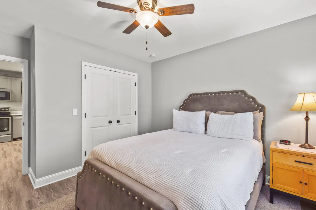 A light-filled bedroom featuring a plush queen bed with luxurious bedding, a nightstand with a lamp, and a ceiling fan overhead. The room exudes a serene and comfortable atmosphere, with natural light streaming in to create a peaceful retreat for relaxation or restful sleep.