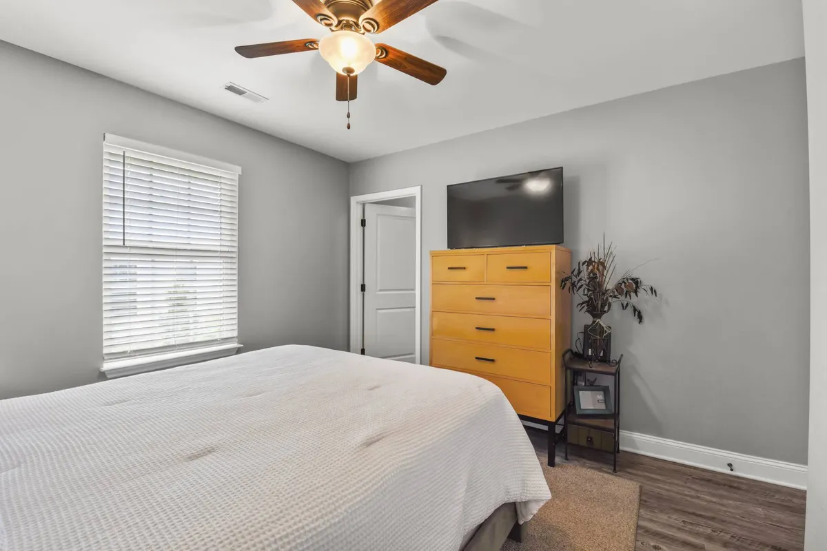 A cozy bedroom with a well-made bed, a bright yellow dresser, a ceiling fan, and a mounted TV. Natural light enters through windows, and the room feels tranquil, inviting, and designed for relaxation or quiet personal time.