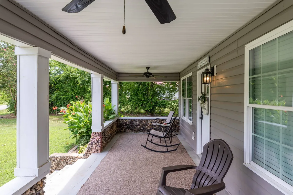 A peaceful exterior view focusing on the front porch area with rocking chairs, a stone wall, and lush green plants. The setting is ideal for enjoying the outdoors, reading, or relaxing while soaking in the sounds of nature.
