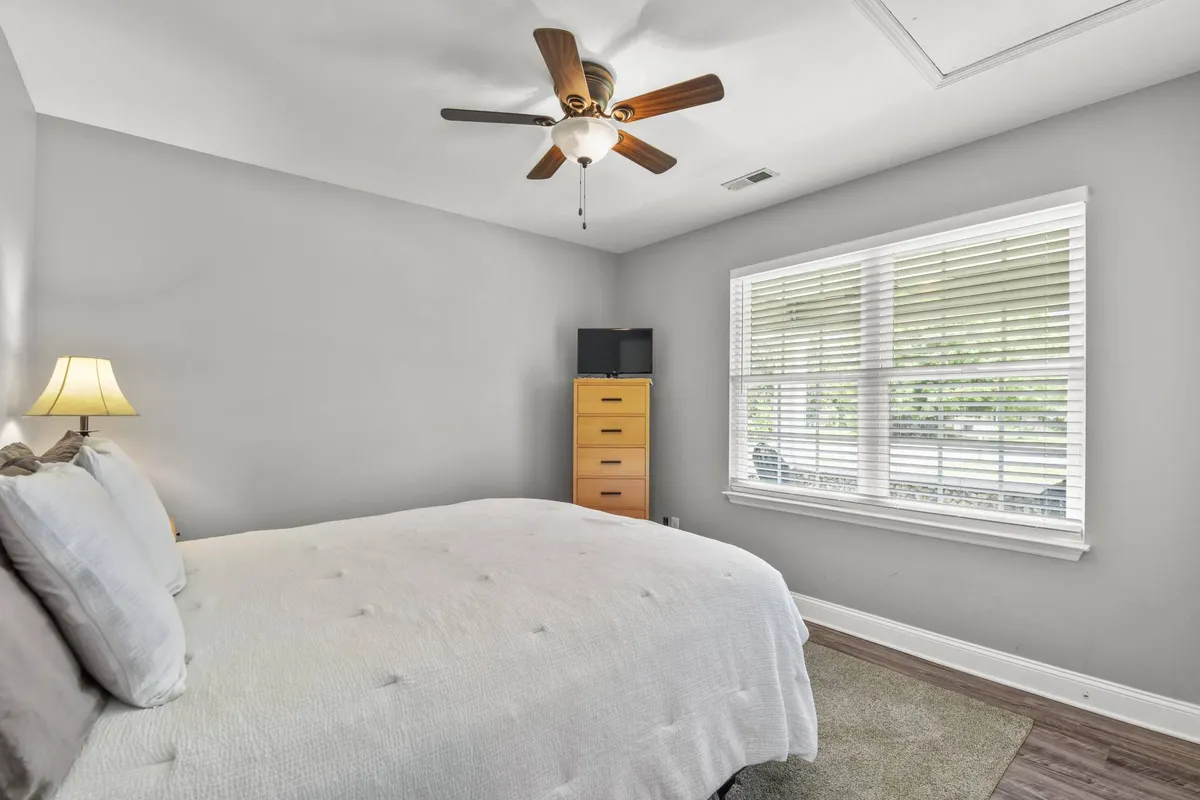 A serene bedroom with a neatly made bed positioned by a window that lets in natural light. A ceiling fan hangs overhead, and a dresser sits against the wall, creating a comfortable and inviting space. The mood is tranquil, perfect for relaxing or enjoying a quiet cup of coffee as sunlight streams in.