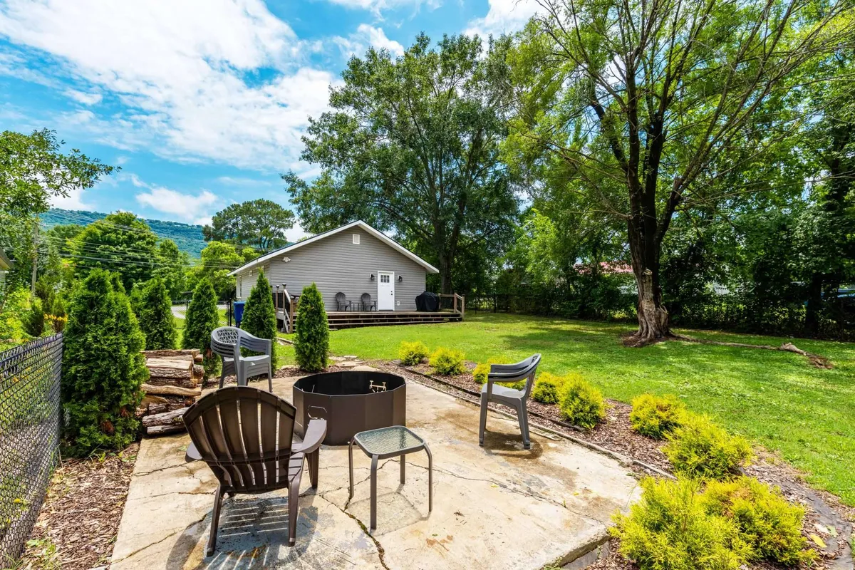 The backyard is shown with a well-maintained green lawn, a cozy fire pit surrounded by outdoor seating, and a glimpse of the house. The scene is set for social gatherings, barbecues, and relaxed evenings, reinforcing the family- and dog-friendly aspect.